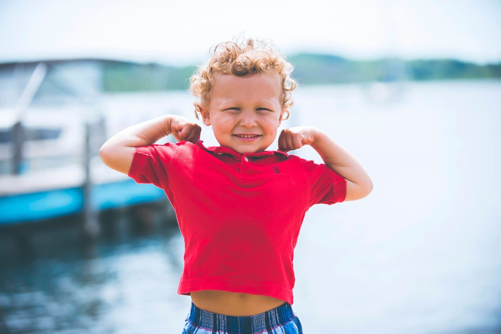 A curly-haired boy flexing his arms and grinning by a lake