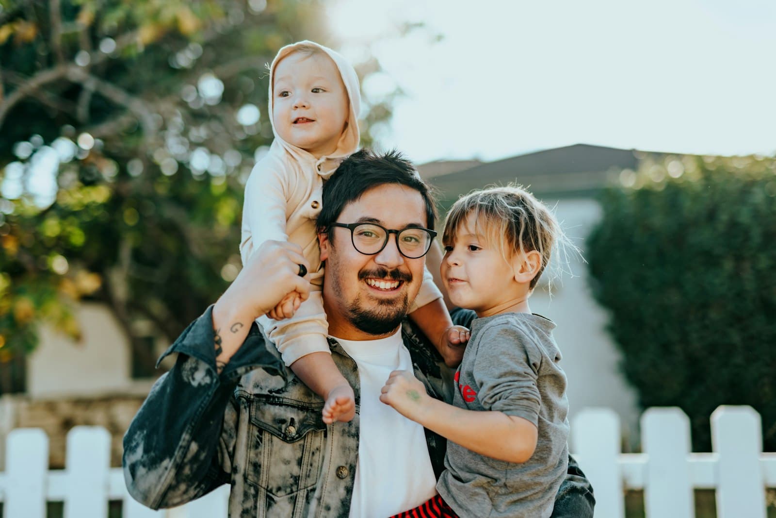 A caregiver carrying two happy children outside a home with a white picket fence